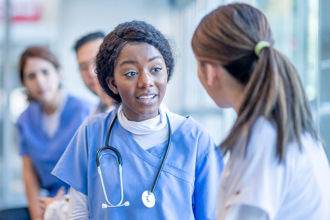 A female doctor of ethnic background is in discussion with someone whilst being overheard by two colleagues