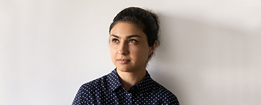 Woman with dark hair pulled back, wearing a navy blue patterned shirt, looking to one side against a plain background