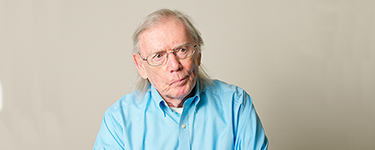 Older man with grey hair and glasses wearing a light blue shirt, standing against a plain background.
