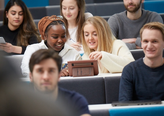 Two students in a lecture discuss something on a tablet