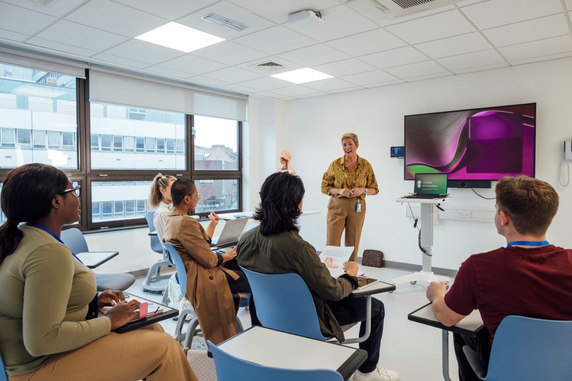 Doctors sat at desks in a classroom environment with a teacher at the front of the classroom presenting